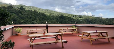 Una terraza en la azotea con mesas de picnic de madera y plantas en macetas, con vistas a una verde ladera.