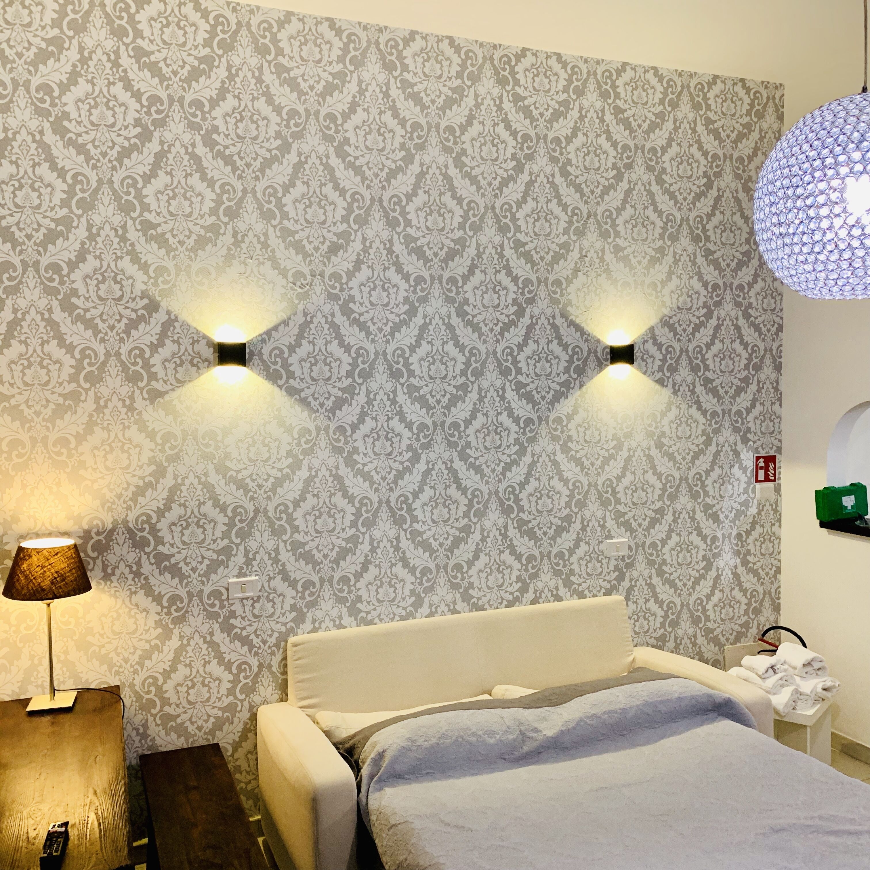 Ein Hotelzimmer mit gemustertem Wandpapier, einem Bett mit weißem Kopfteil, zwei Wandleuchten, einer Stehlampe und einem grünen Notfallkasten an der Wand.