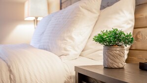 A neatly made bed with a white comforter and pillows, a bedside table with a potted plant, and a lamp in the background. - Stay Avenida Westshore (Tampa)