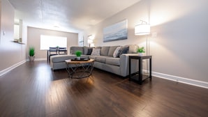 A modern living room with hardwood flooring, a sectional sofa, a coffee table, and a side table with a lamp. - Stay Avenida Westshore (Tampa)