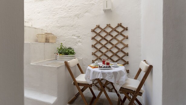 Ein kleiner Essbereich im Freien mit einem weißen Tisch und Stühlen, einer Spalierwand und einer kleinen Pflanze auf einem Regal.