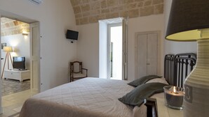 Ein Schlafzimmer mit einem großen Bett, einem an der Wand montierten Fernseher, einem Sessel, einer Tür zu einem anderen Raum und einem Fenster mit Vorhängen.