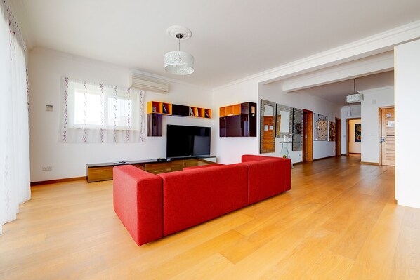 A spacious living room with a red sofa, a flat-screen TV, and a wooden floor. - Seafront Apartment with Pool, Upmarket Complex (Sliema)