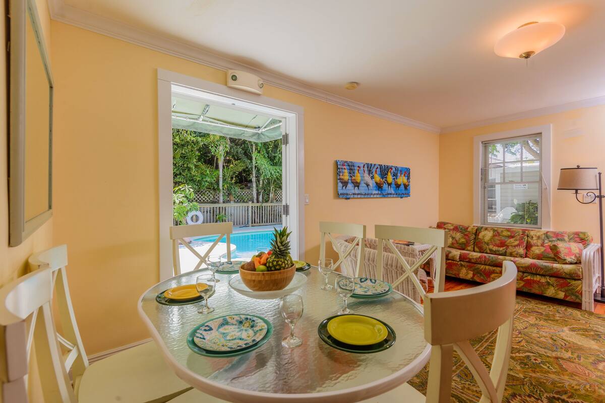 A dining area with a glass table set for four, surrounded by white chairs. A bowl of fruit is on the table, and there is a view of a pool through an open door.