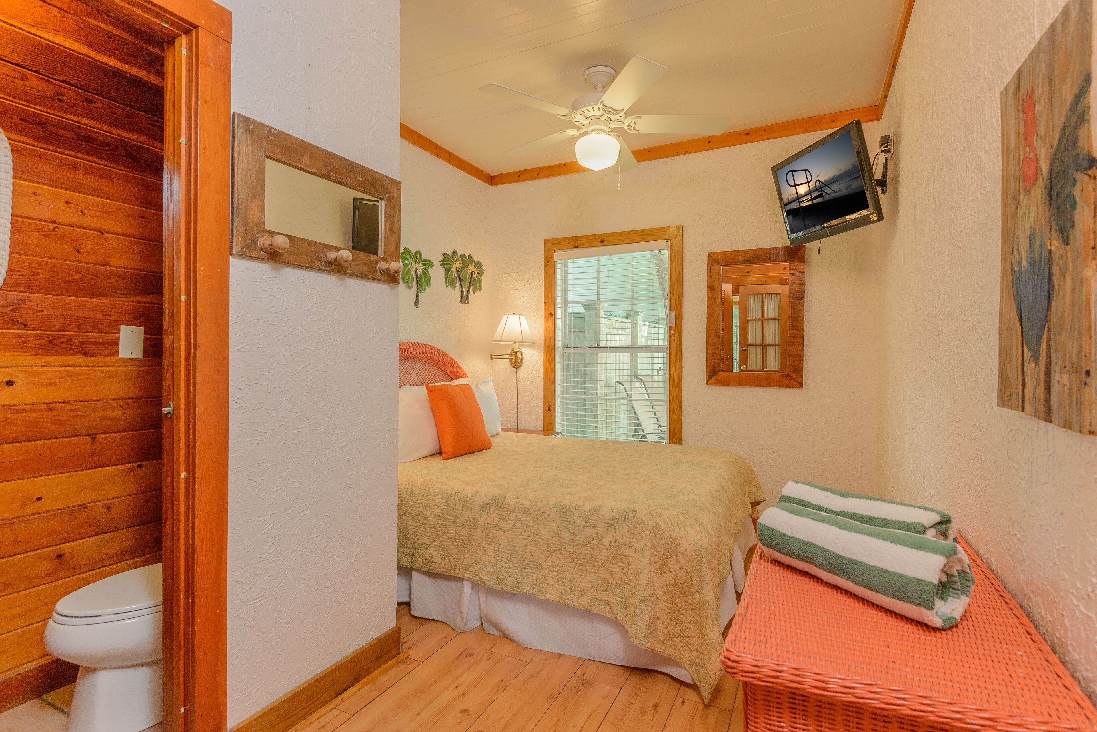 A small, cozy room with a bed, a wooden door, a ceiling fan, a window with blinds, and a wall-mounted TV.