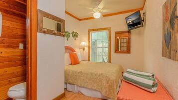 A small, cozy room with a bed, a wooden door, a ceiling fan, a window with blinds, and a wall-mounted TV.