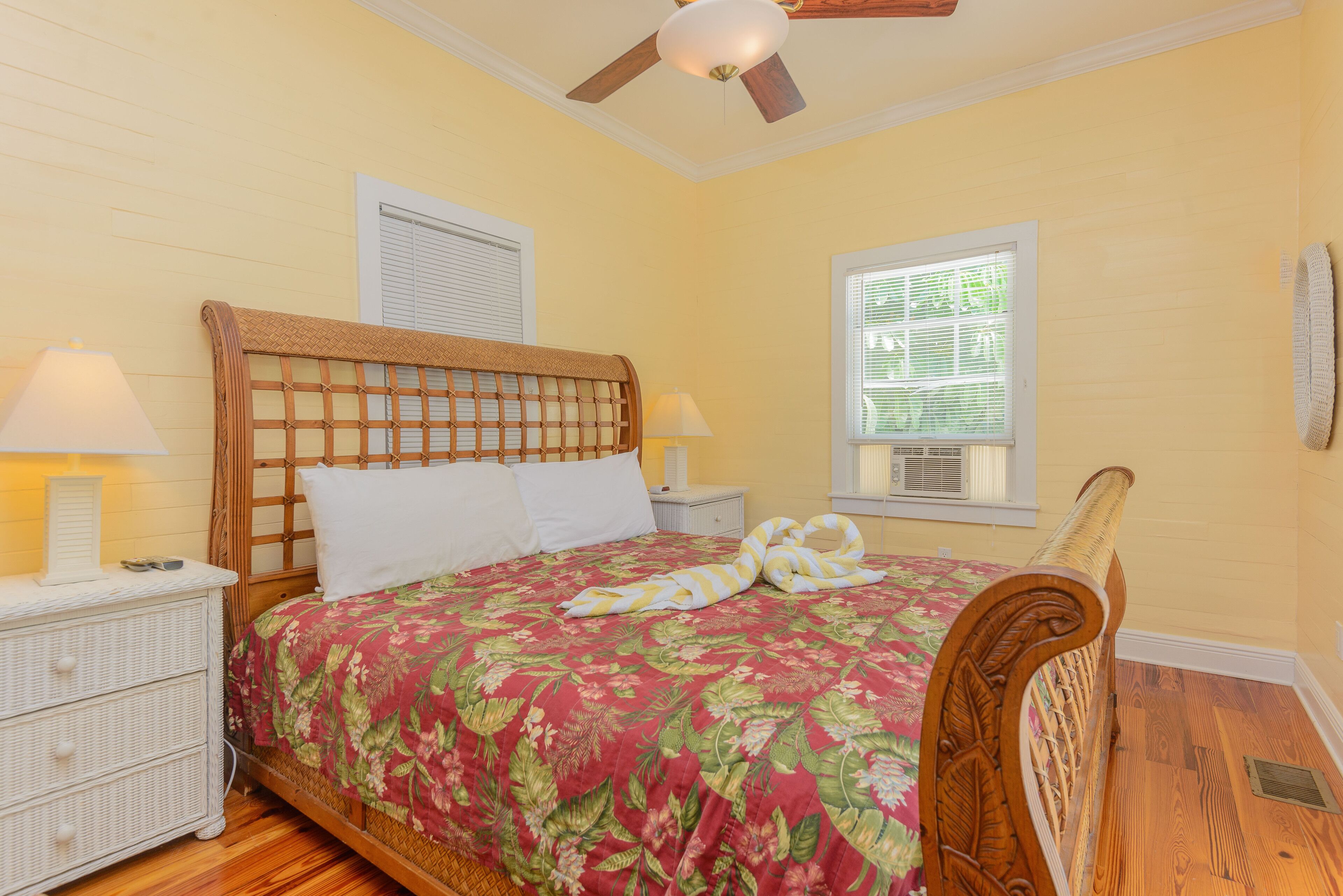 A bedroom with a wooden headboard, a floral bedspread, a bedside table with a lamp, and a window with blinds.