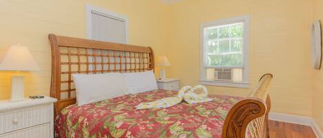 A bedroom with a wooden headboard, a floral bedspread, a bedside table with a lamp, and a window with blinds.