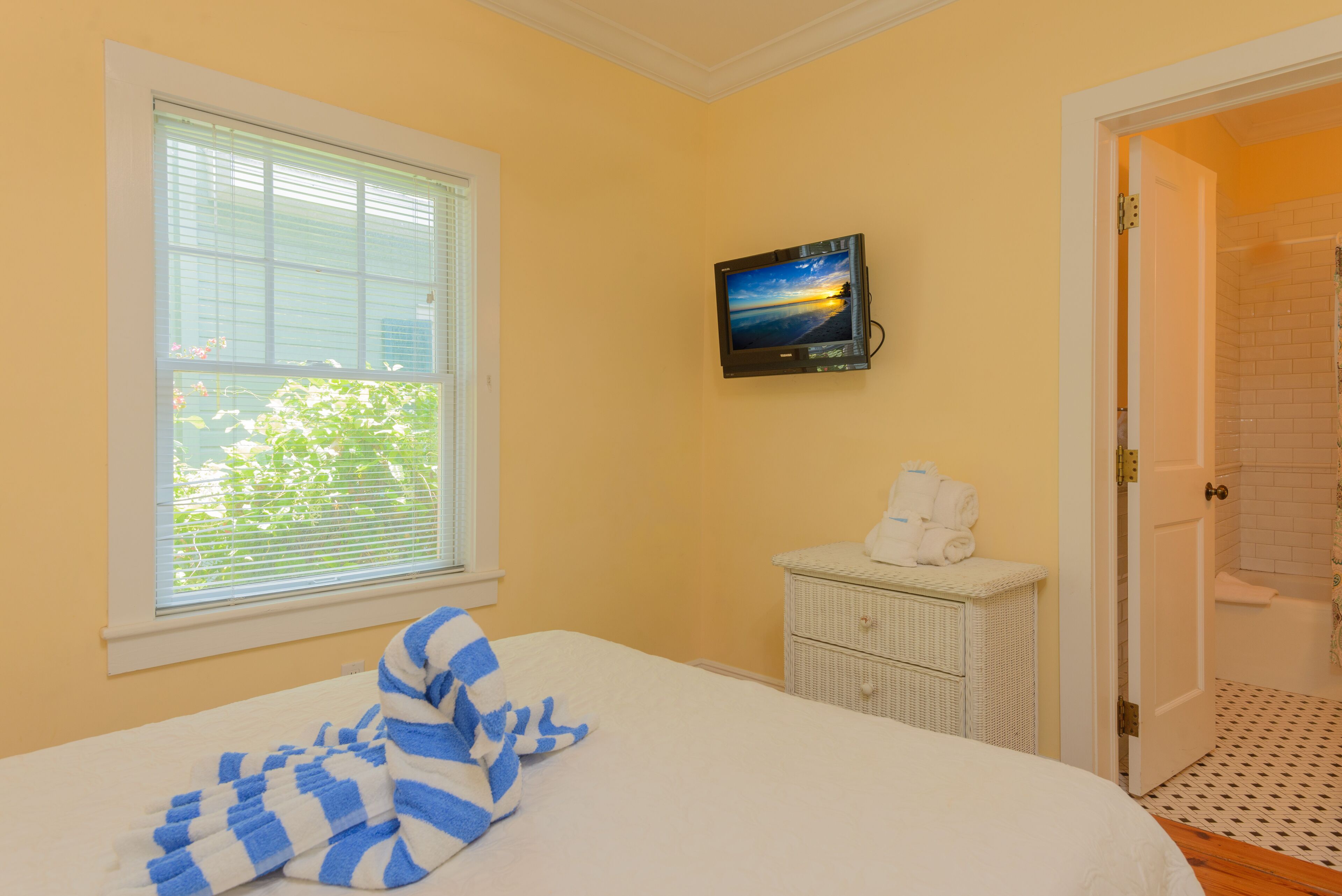 A bedroom with a bed, a window with blinds, a television on the wall, a small white dresser, and a bathroom door.