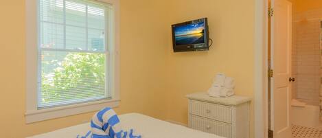 A bedroom with a bed, a window with blinds, a television on the wall, a small white dresser, and a bathroom door.