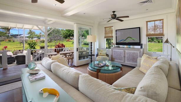 Un salon spacieux avec un grand canapé, une table basse en verre et un téléviseur. Il y a un balcon avec des meubles d’extérieur et une vue sur un jardin.