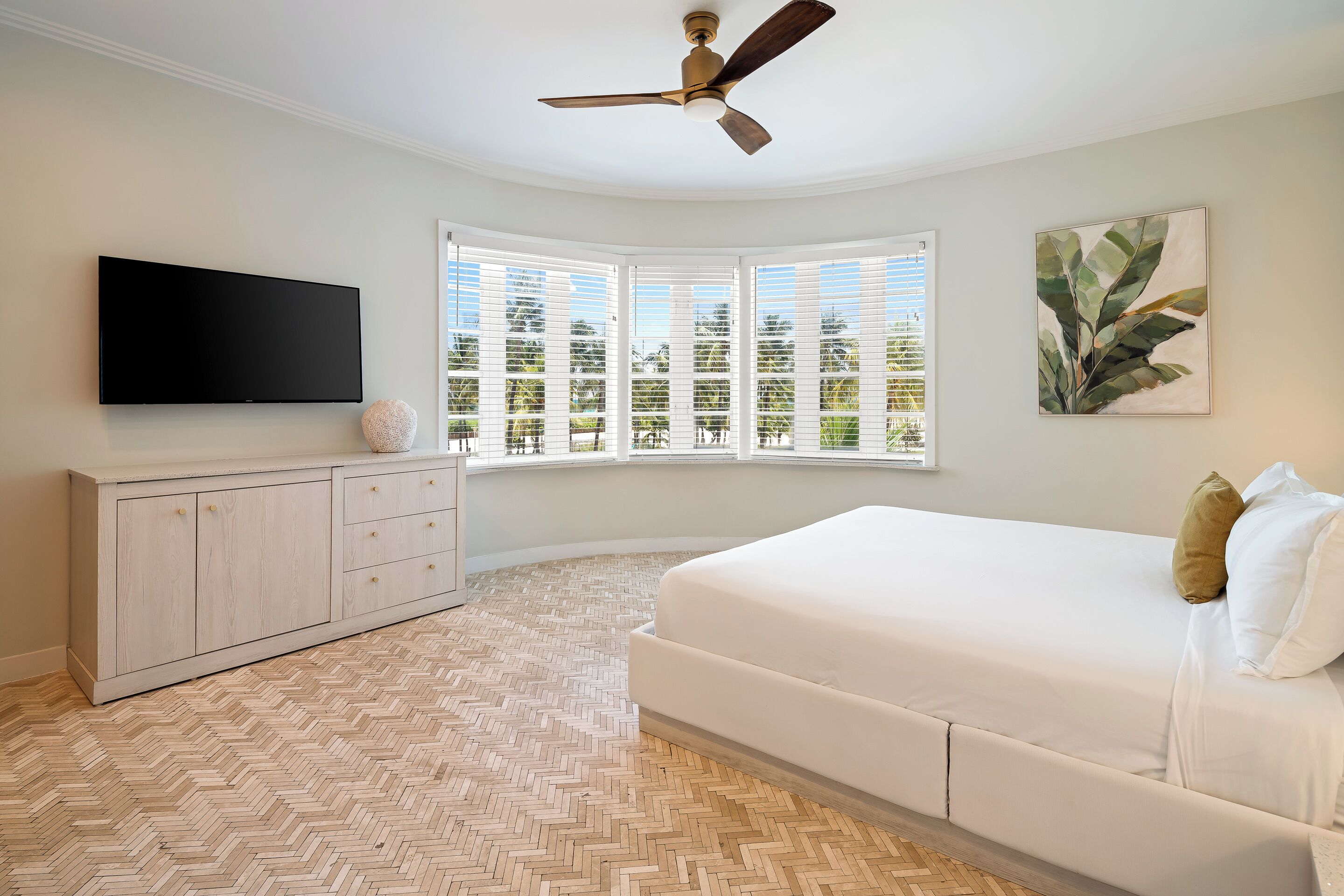 A bedroom with a large bed, a flat-screen TV, a ceiling fan, a herringbone-patterned floor, a window with blinds, and a wall-mounted artwork.