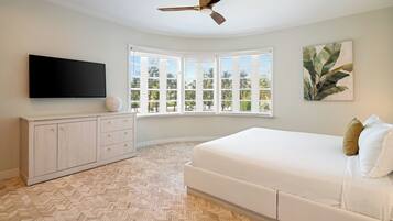 A bedroom with a large bed, a flat-screen TV, a ceiling fan, a herringbone-patterned floor, a window with blinds, and a wall-mounted artwork.