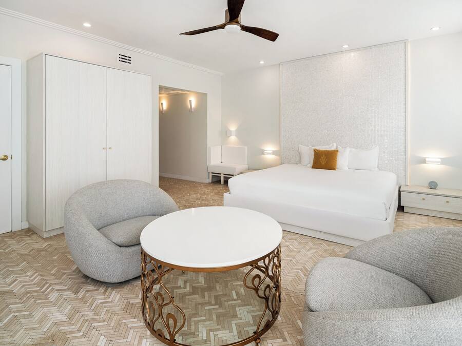 Quarto de hotel moderno com uma cama grande, duas poltronas cinzentas, uma mesa redonda e um ventilador de teto.