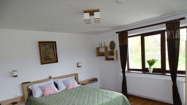 Un dormitorio con cama, cabecero de madera, mesitas de noche, ventana con cortinas, radiador y estante con objetos decorativos.