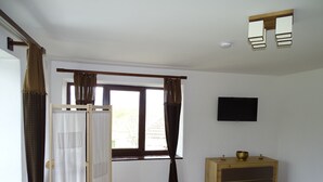 Una habitación con piso de madera, una ventana con cortinas, un televisor montado en la pared y un mueble de madera con cajones.