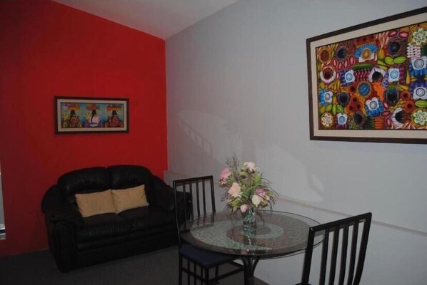 A room with a black leather sofa, a glass table with chairs, a vase of flowers, and two framed artworks on the walls. - Apart Hotel Valle Verde (San Salvador)