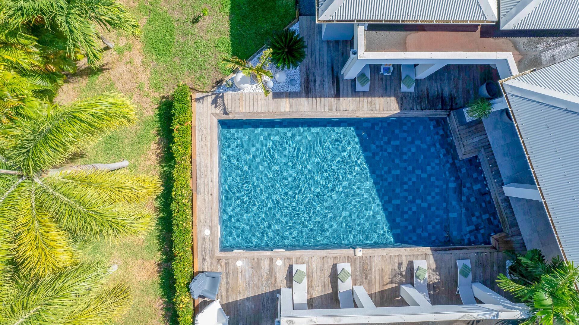An aerial view of a rectangular swimming pool with a wooden deck surrounding it, adjacent to a house with a sloped roof.