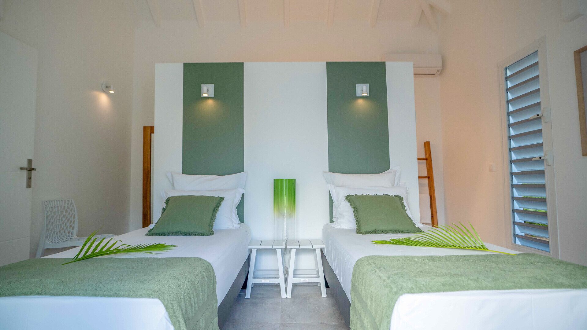 A room with two beds, each with a green pillow and a white bedspread. There is a small table between the beds, and a window with blinds on the right side.
