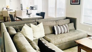 A modern living room with a grey sectional sofa, a wooden coffee table, and a dining area in the background.