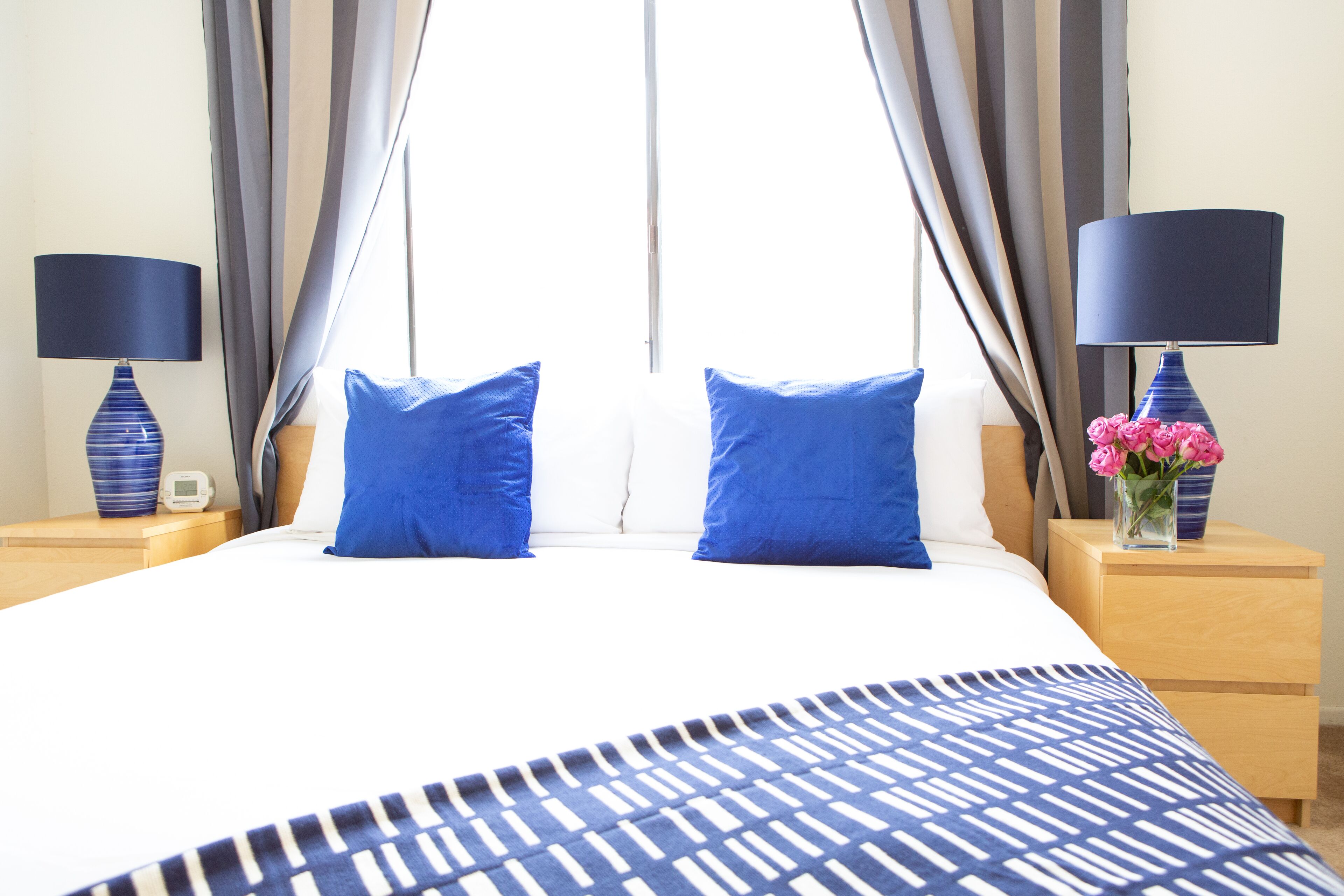 A hotel room with a large bed, two blue pillows, a patterned bedspread, a nightstand with a lamp, and a vase with pink flowers.