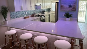 A modern kitchen with white cabinets, a large island with bar stools, and a painting of a beach scene. - Direct Gulf View, Beach Condo on Manasota Key, FL (Englewood)