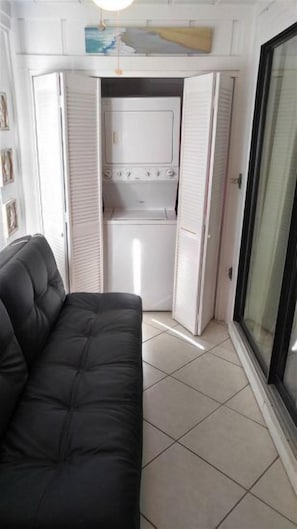 Een kamer met een zwarte bank, een wit wasgedeelte met een droger, en een glazen deur.