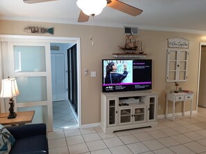 Una sala de estar con televisor, ventilador de techo, mueble y puerta que da a otra habitación.