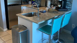 Eine moderne Küche mit einem Edelstahl-Kühlschrank, einer Mikrowelle, einer Kaffeemaschine und einem Spülbecken. Zwei blaue Barhocker und ein Mülleimer sind ebenfalls vorhanden.