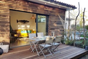 Un espace de repas extérieur moderne, doté d’une terrasse en bois, d’une table dressée pour deux personnes et d’une porte vitrée donnant sur un espace intérieur avec un canapé jaune et une fresque représentant une baleine.