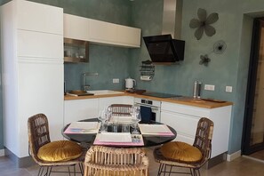 Une cuisine compacte avec des meubles blancs, une cuisinière noire et un coin repas composé de chaises en osier et d’une table en verre, le tout disposé pour accueillir quatre personnes.