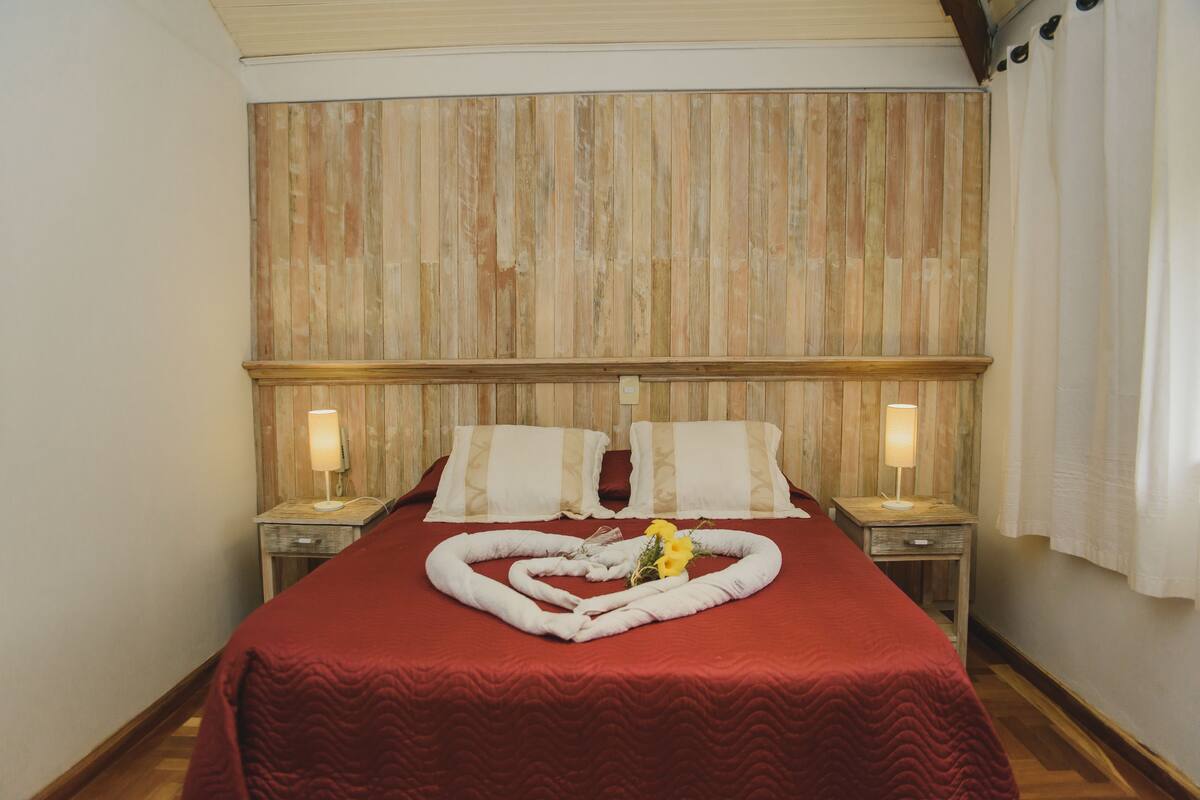 A hotel room with a wooden headboard, a red bedspread, two bedside tables with lamps, and a heart-shaped towel arrangement on the bed.