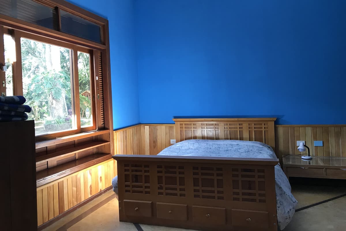A bedroom with a wooden bed, a window with a view of greenery, and a bedside table with a lamp.