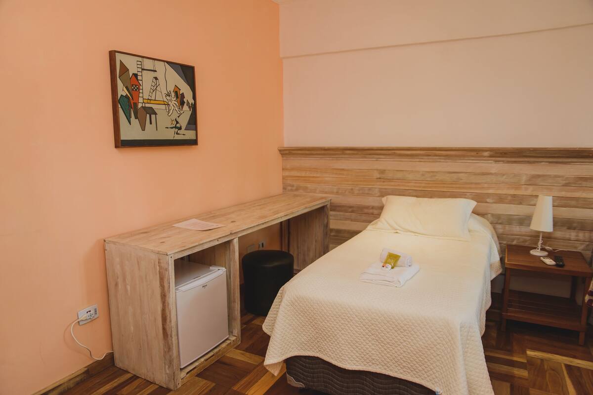 A room with a bed, a desk, a small refrigerator, and a painting on the wall.