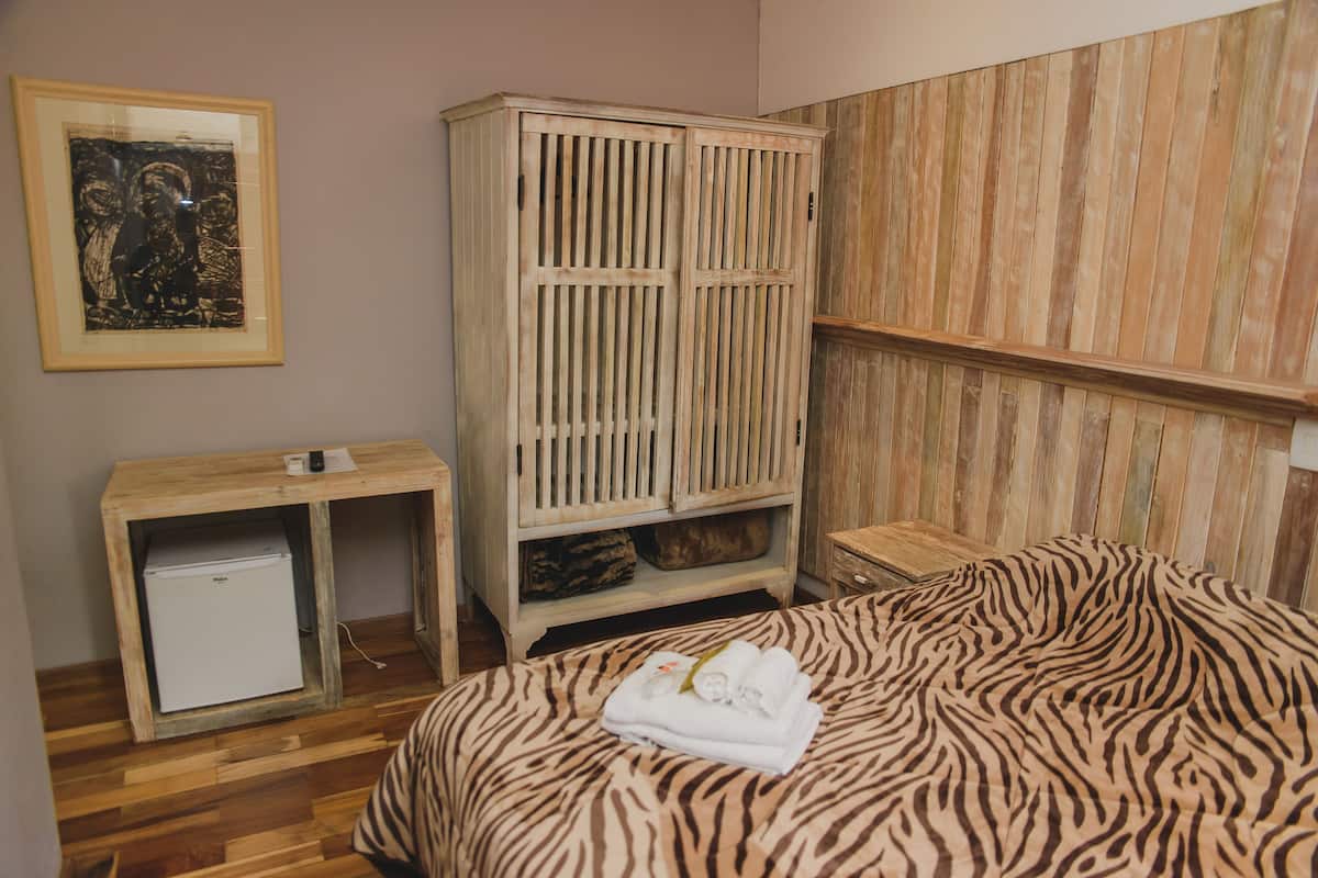 A bedroom with a bed, a wooden wardrobe, a small desk with a refrigerator, and a wooden panel wall.