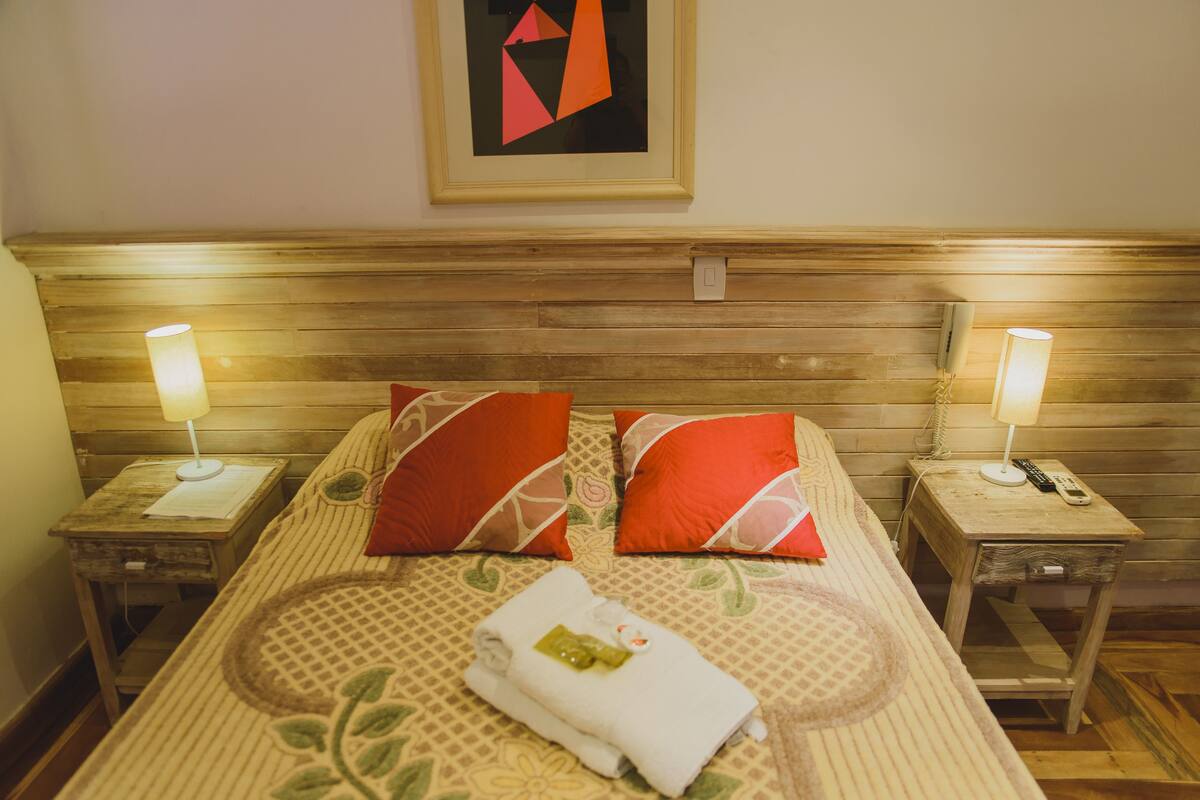 A neatly made bed with two red pillows, a white towel, and a framed abstract painting on the wall.