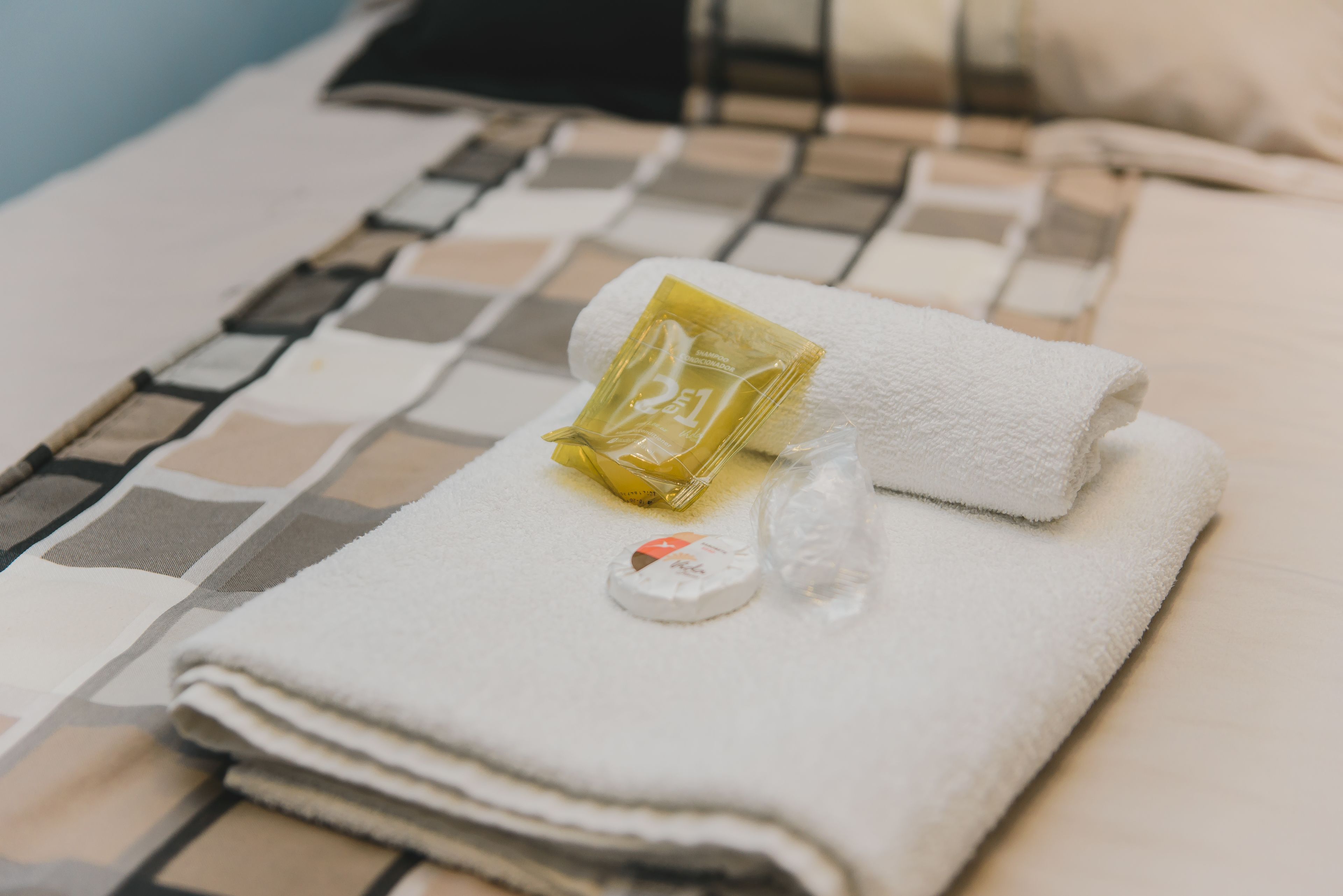 A neatly folded white towel with a small bottle and a packet on a checkered bedspread.