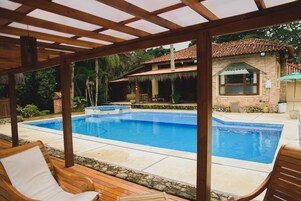 Uma área de piscina coberta com espreguiçadeiras de madeira, um caminho de pedra e vista para uma casa com telhado em cerâmica.