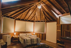 Quarto com teto de madeira inclinado, cama com colcha listrada, mesa de cabeceira com abajur e um pequeno frigorífico.