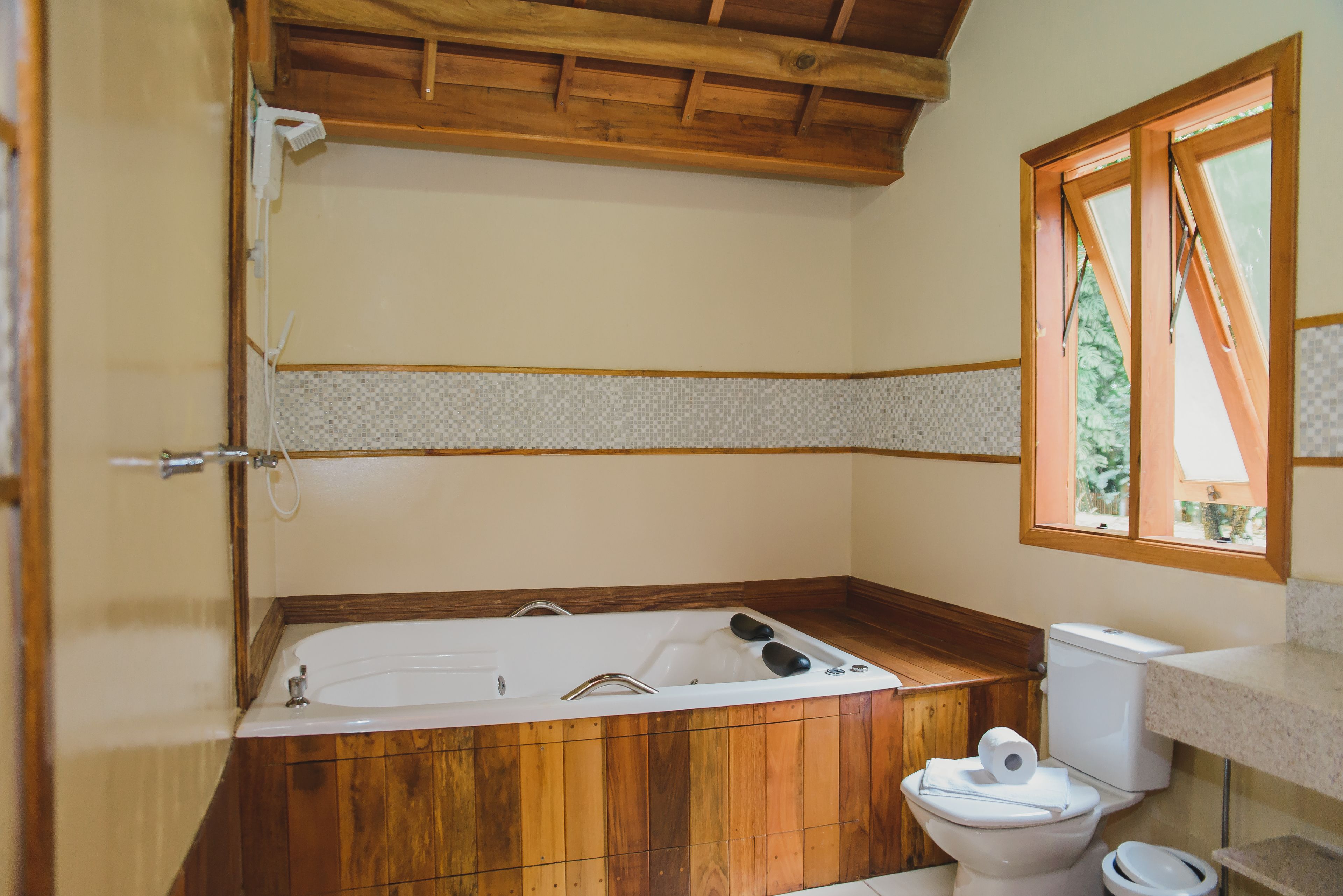 A bathroom with a wooden bathtub, a toilet, and a window with a view of trees.