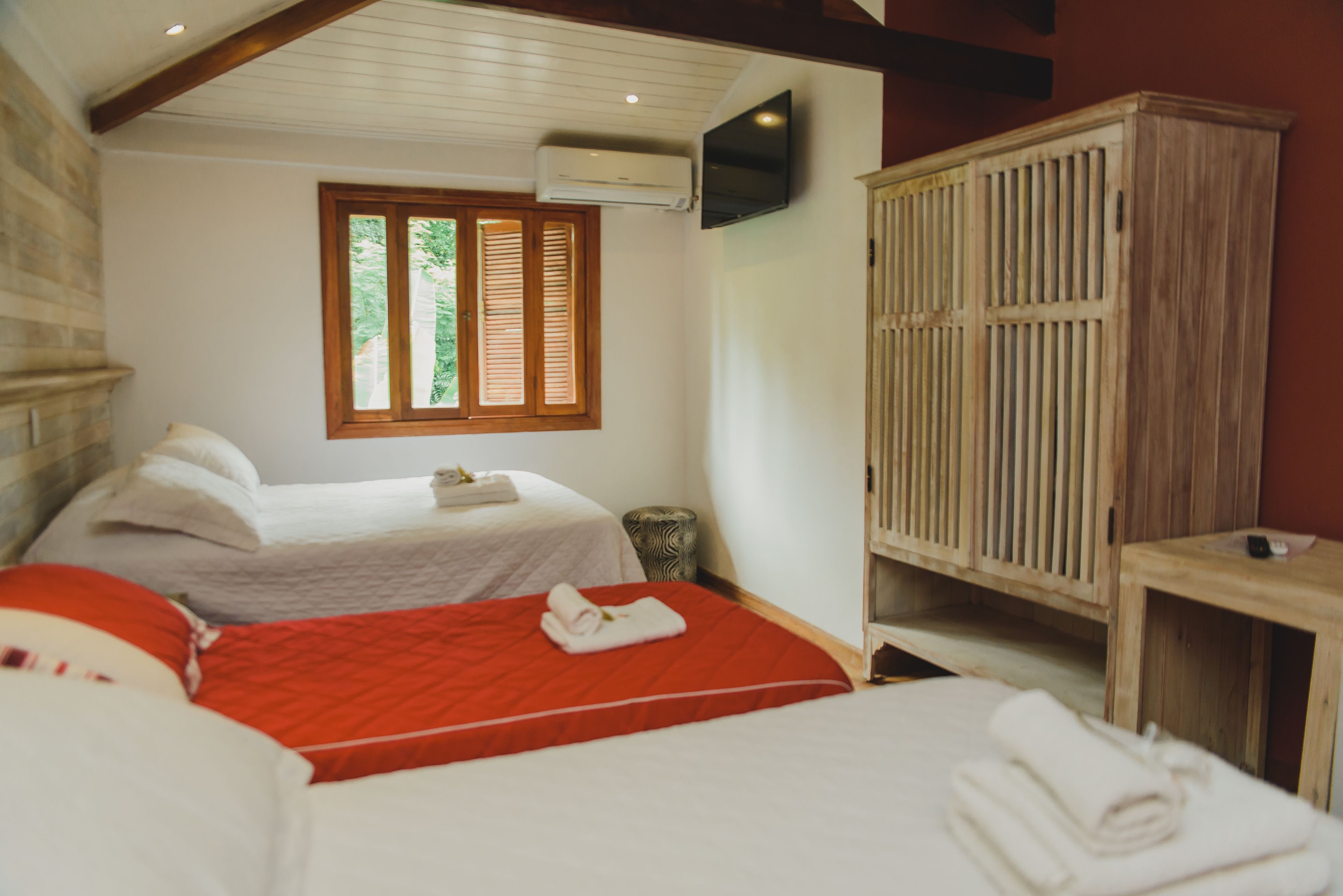 Una habitación con dos camas, una con colcha roja y la otra con colcha blanca. Hay un armario de madera y un televisor montado en la pared.