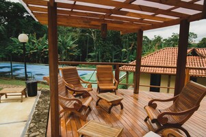 Um deck de madeira com cadeiras de descanso e mesas, com vista para uma piscina e vegetação exuberante.