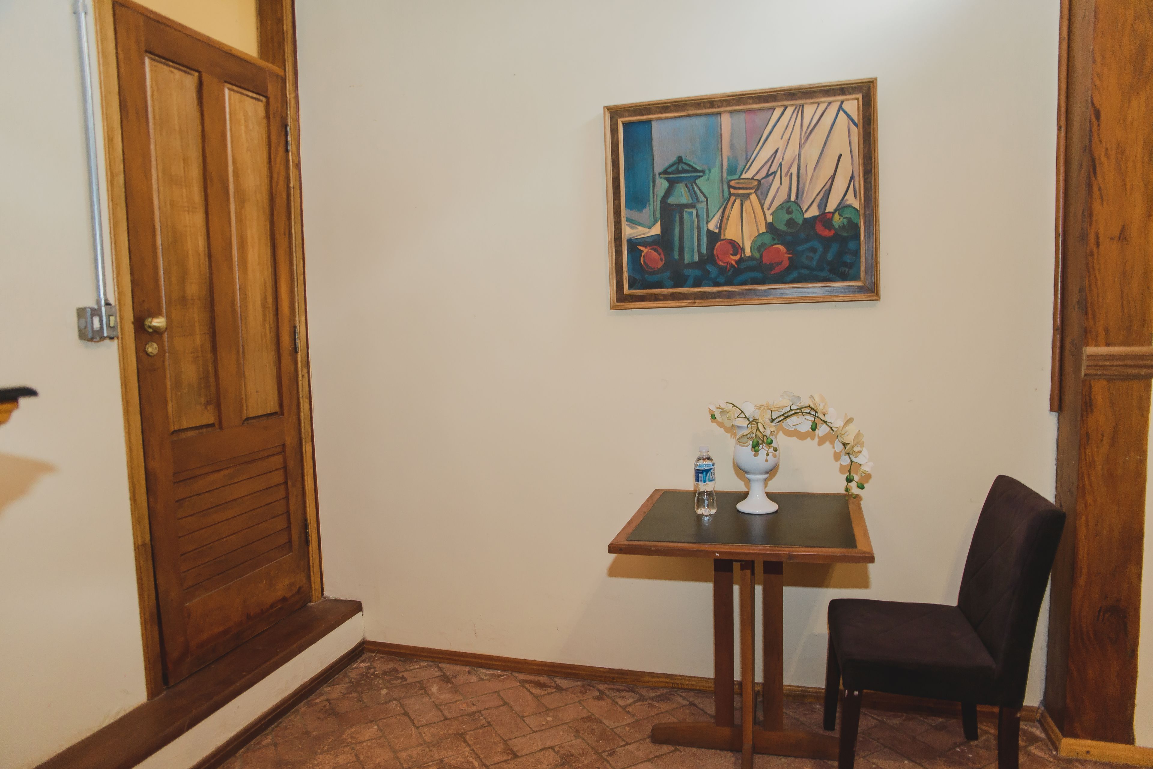 A hallway with a wooden door, a framed painting, a small table with a vase and flowers, and a chair.