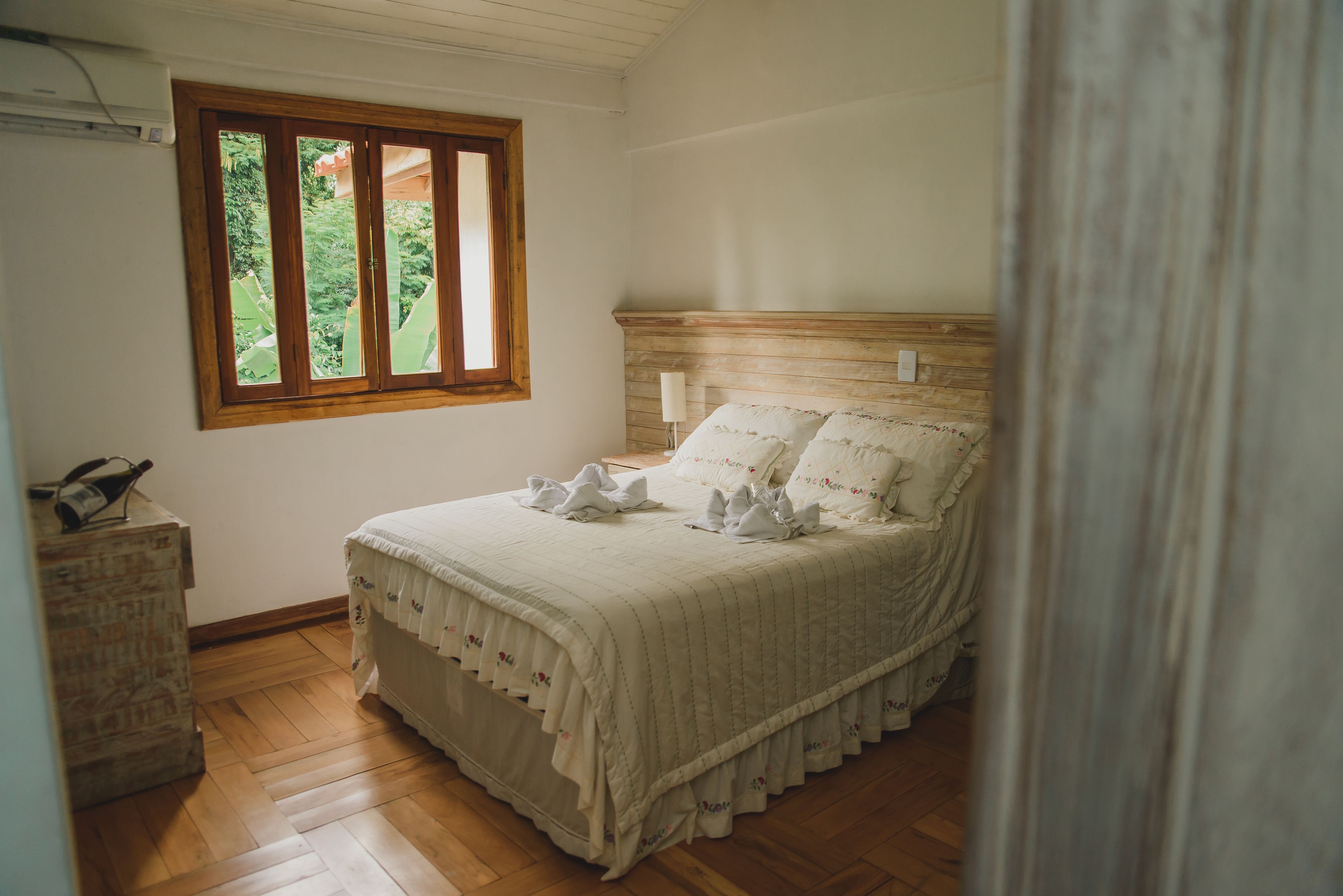 Un dormitorio con cabecera de madera, ventana con vista a la vegetación, piso de madera y mesita de noche con lámpara.