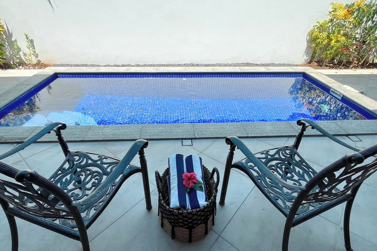 Ein Pool mit blauen Mosaikfliesen, umgeben von einer gefliesten Terrasse mit zwei verzierten Metallstühlen und einem Korbstuhl mit gestreiftem Sitzkissen.