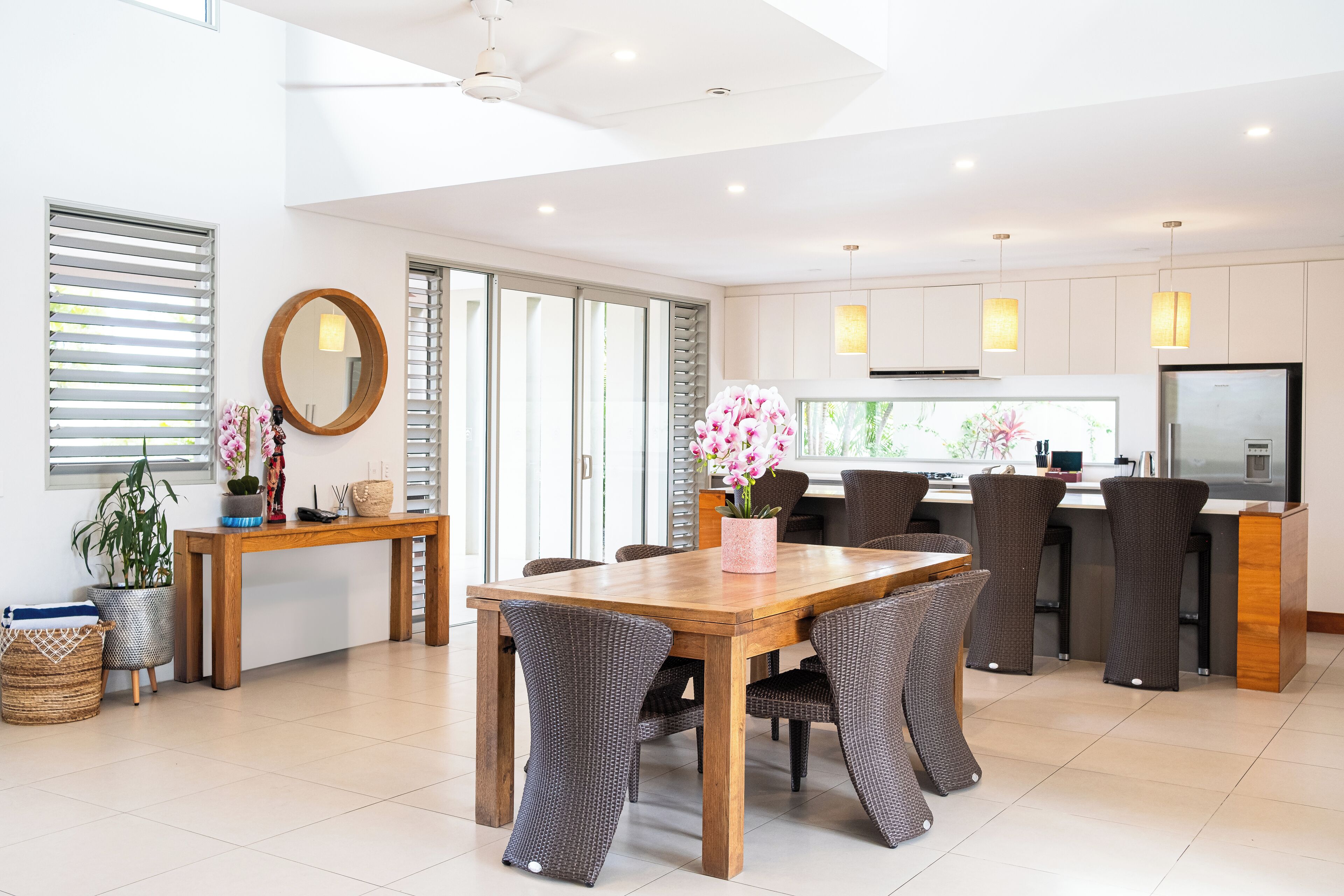 A modern dining area with a wooden table and wicker chairs, a kitchen with a large island, and a view of the outdoors through the sliding doors.