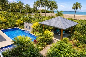 Ein Poolbereich mit Steinmauer, umgeben von tropischen Pflanzen, einem Pavillon und Blick auf den Strand.