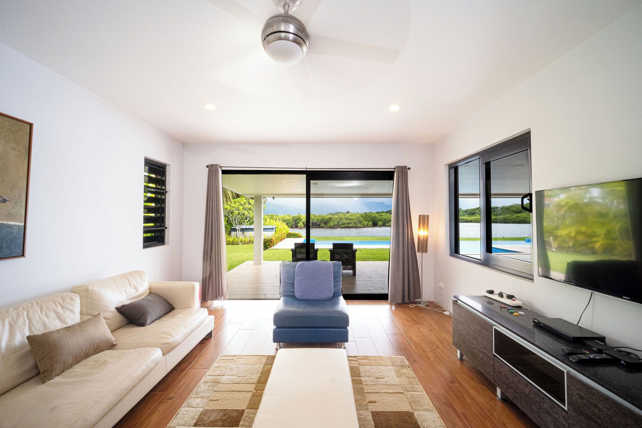 A modern living room with a sofa, a blue chair, a TV, and a view of a pool and greenery.