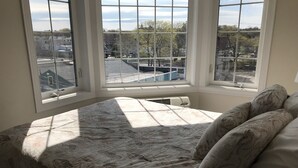 A bedroom with a large bed, a window with multiple panes, and a view of buildings and trees outside. - Atlantic Ocean Suites - Sand Castle Suite 7 (Old Orchard Beach)