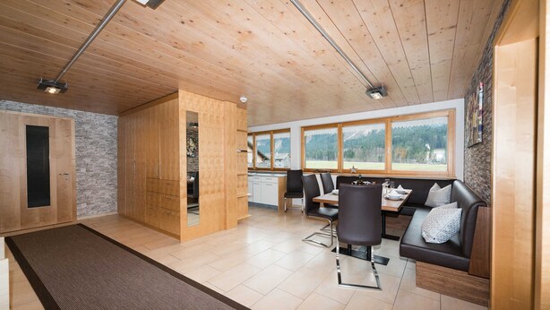A spacious room with wooden ceiling and walls, a dining area with a table and chairs, a sofa, and a view of the outdoors through large windows.